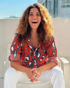 Fotografia de uma mulher sorridente com cabelos cacheados castanhos, vestindo camisa vermelha com estampas abstratas e calça branca, sentada sob luz solar.