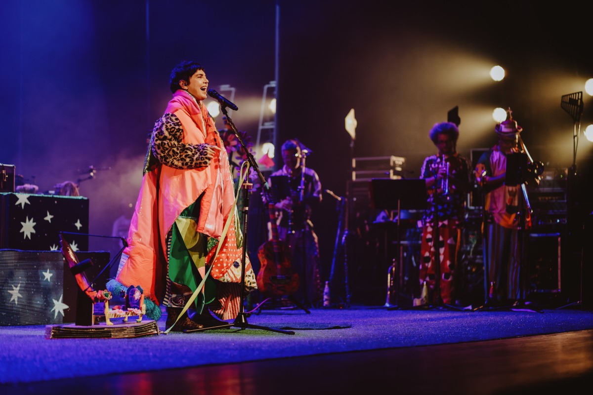 Cantora vestida com roupas coloridas e casaco de estampa animal canta ao microfone em palco iluminado, acompanhada por banda com instrumentos de sopro ao fundo.