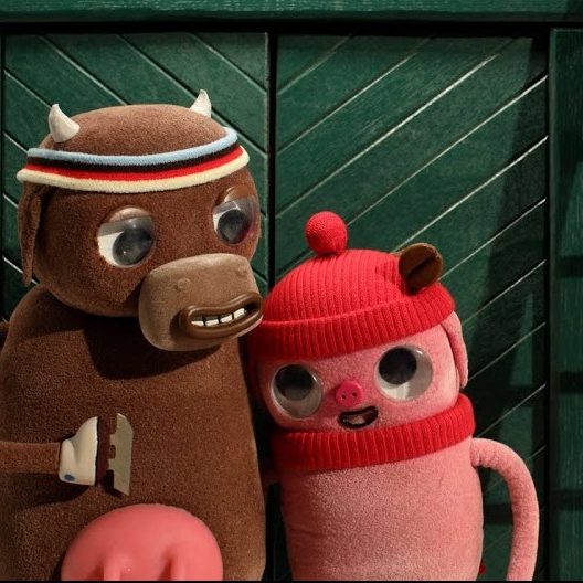 Imagem de dois bonecos de pelúcia: um touro marrom abraça um porquinho rosa de gorro vermelho diante de uma porta verde.