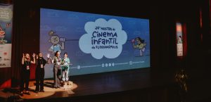 Quatro mulheres no palco de um teatro apresentam-se diante de uma grande tela que exibe o título “Vigésima quarta Mostra de Cinema Infantil de Florianópolis”, com ilustrações coloridas de personagens infantis.
