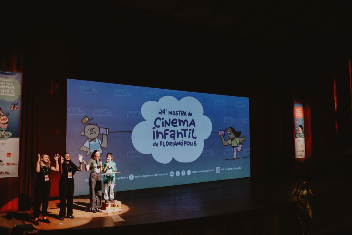 Quatro mulheres no palco de um teatro apresentam-se diante de uma grande tela que exibe o título “Vigésima quarta Mostra de Cinema Infantil de Florianópolis