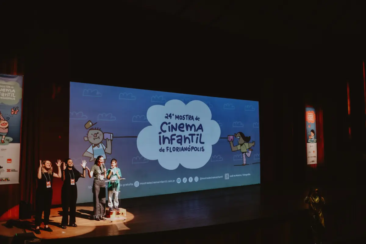 Quatro mulheres no palco de um teatro apresentam-se diante de uma grande tela que exibe o título “Vigésima quarta Mostra de Cinema Infantil de Florianópolis