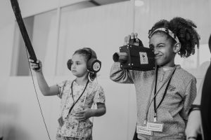 menina e instrutor manuseiam câmera e microfone em oficina audiovisual.