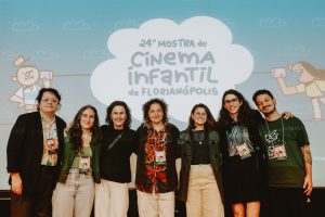 grupo de mulheres posa sorridente diante do painel azul da Mostra de Cinema Infantil de Florianópolis.