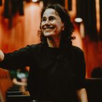 mulher de preto sorri e acena em ambiente de teatro.