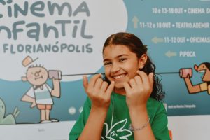 menina de camiseta verde sorri em frente ao painel azul do evento.