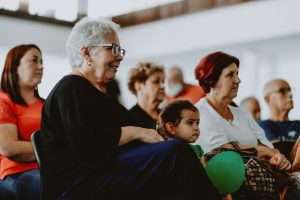 grupo de pessoas sentadas, incluindo idosos e crianças, assiste concentrado a uma atividade.