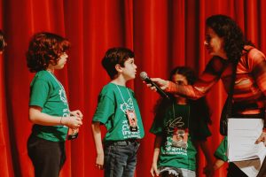 crianças e mulher em frente a cortina vermelha participam de oficina de cinema.