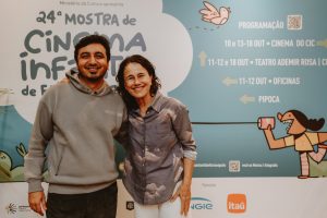 homem e mulher posam sorridentes diante de painel azul da Mostra de Cinema Infantil de Florianópolis.