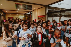 multidão de crianças em fila no saguão de um cinema, algumas segurando balões.