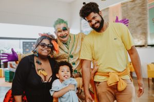 dois artistas caracterizados e sorridentes posam com duas crianças pequenas, uma delas nos braços de um dos artistas.