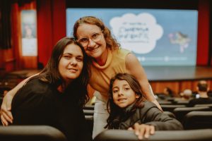 mulher abraça duas crianças sorridentes dentro de uma sala de cinema.