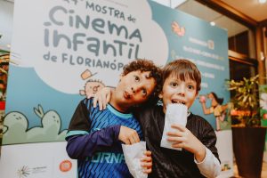 dois meninos posam diante de painel azul da Mostra de Cinema Infantil de Florianópolis, segurando copos de pipoca.
