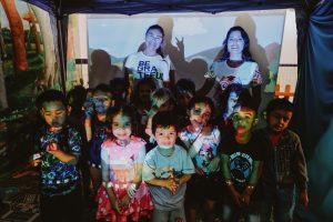 Grupo de crianças sorridentes em frente a uma projeção colorida, acompanhadas por duas mulheres ao fundo, em ambiente escolar decorado com pinturas de árvores.