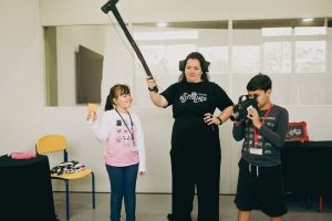 Mulher com camiseta preta segura um microfone boom sobre duas crianças; uma menina sorri segurando uma claquete e um menino grava com uma câmera em uma oficina de cinema.