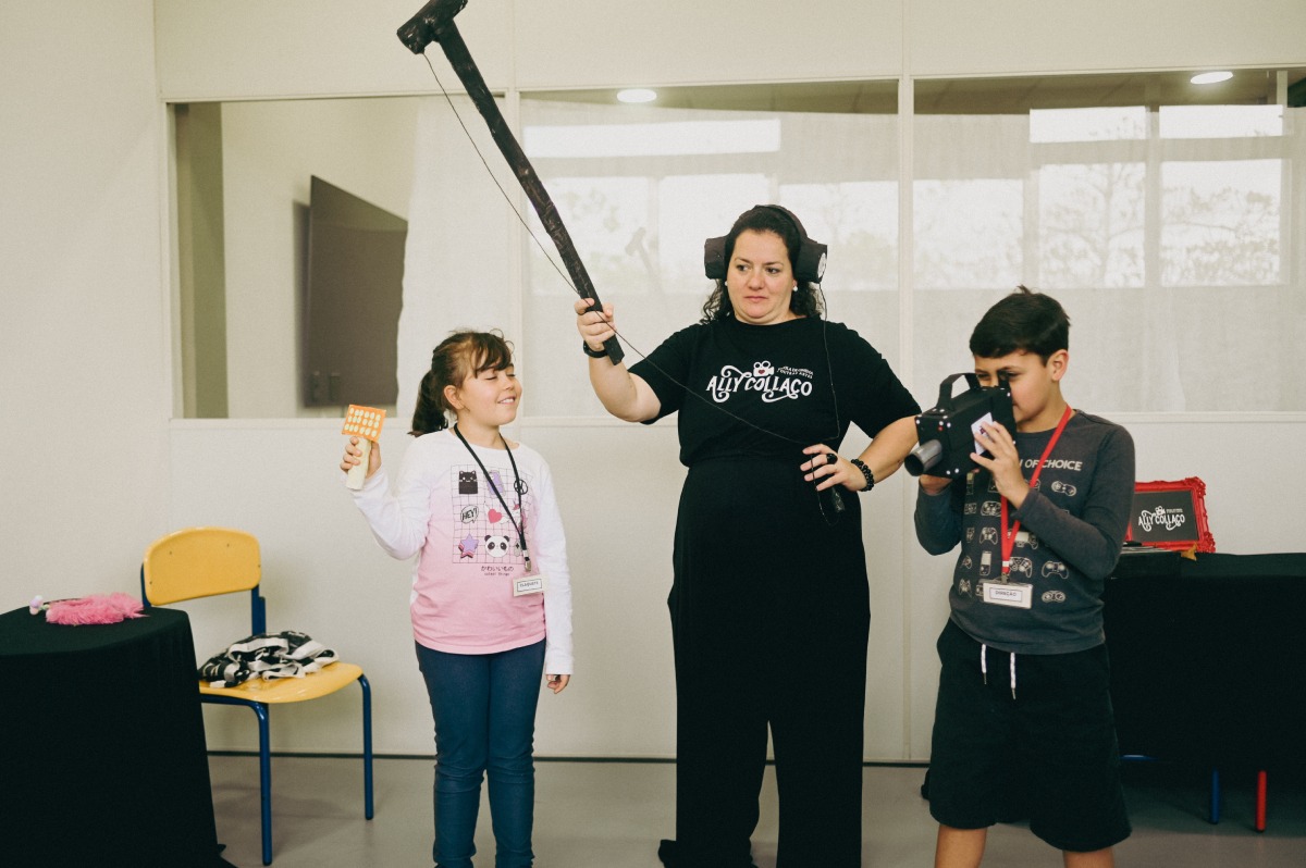 Mulher com camiseta preta segura um microfone boom sobre duas crianças; uma menina sorri segurando uma claquete e um menino grava com uma câmera em uma oficina de cinema.