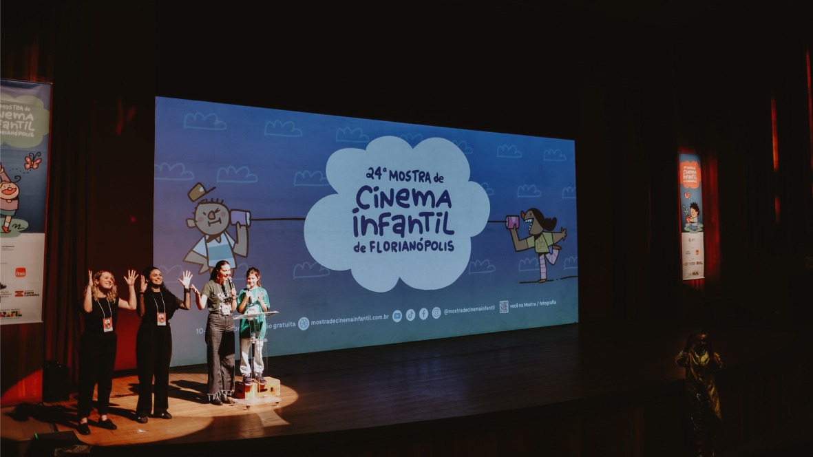 Fotografia de palco com quatro mulheres sob holofote diante de telão com a frase: “Vigésima quarta Mostra de Cinema Infantil de Florianópolis”.