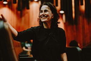 Fotografia de mulher sorridente, de blusa preta, sentada em auditório com paredes de madeira, acenando.