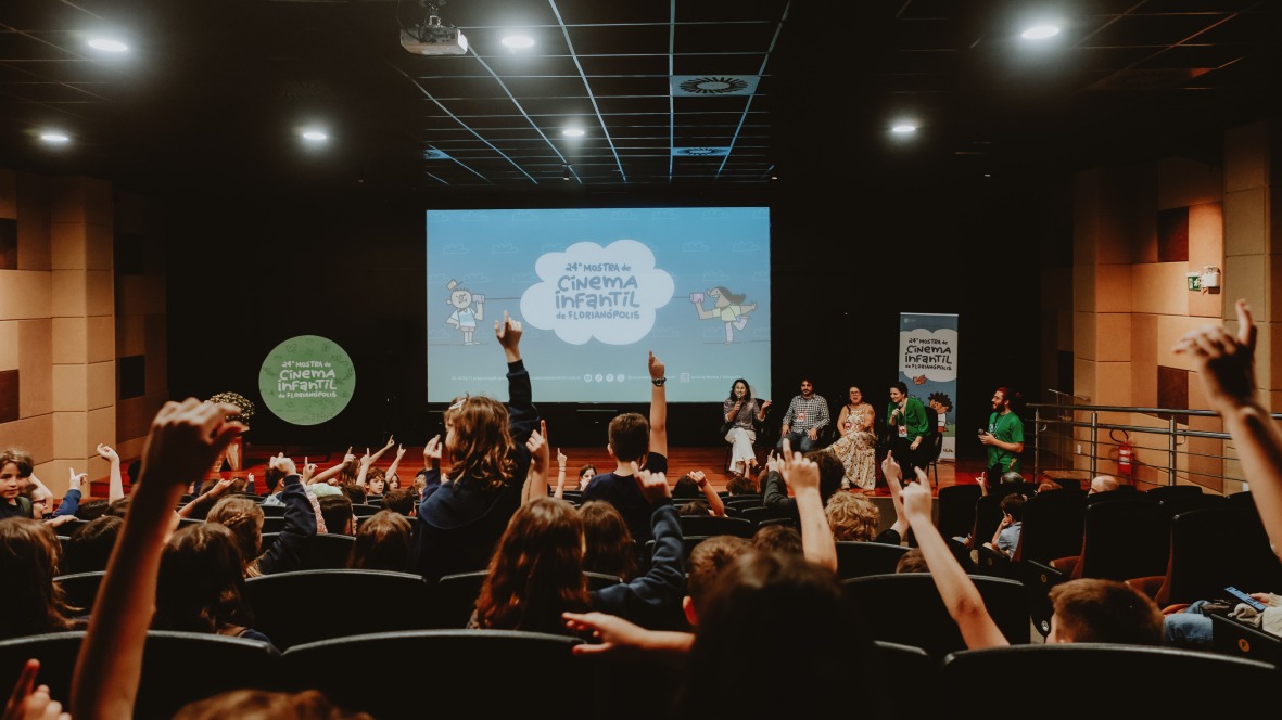 Fotografia de auditório lotado de crianças com braços erguidos diante do palco. No telão, lê-se: “Vigésima quarta Mostra de Cinema Infantil de Florianópolis”.