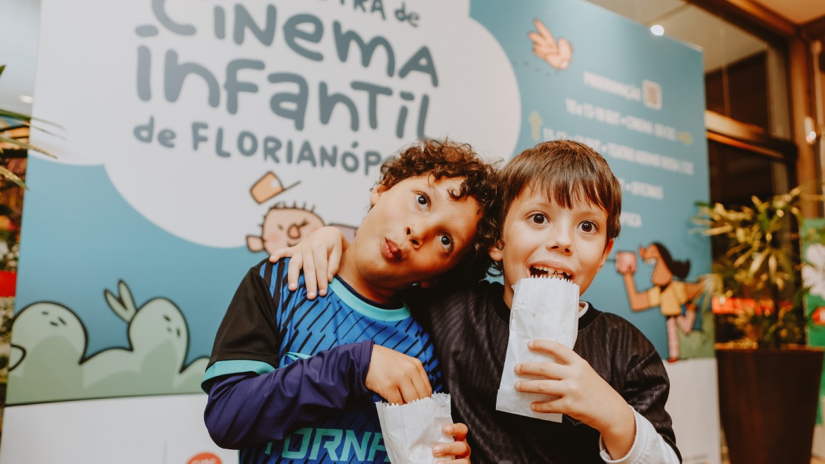 Fotografia de dois meninos abraçados, fazendo caretas e segurando saquinhos de pipoca, diante de painel da Vigésima quarta Mostra de Cinema Infantil de Florianópolis.