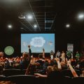 Fotografia de auditório lotado de crianças com braços erguidos diante do palco. No telão, lê-se: “Vigésima quarta Mostra de Cinema Infantil de Florianópolis”.
