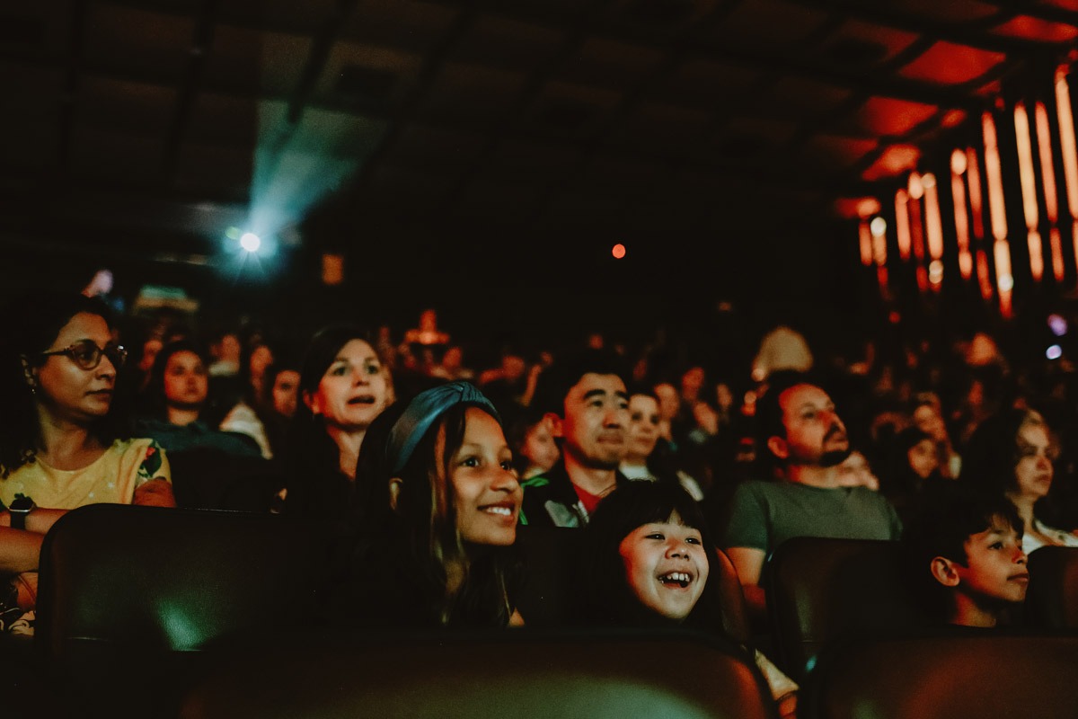 Fotografia de plateia em cinema escuro, com crianças sorrindo e adultos atentos à exibição.