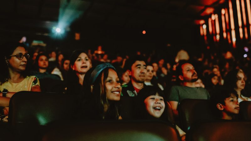 Fotografia de plateia em cinema escuro, com crianças sorrindo e adultos atentos à exibição.