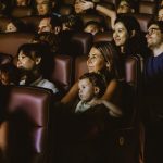 Fotografia de plateia em sala de cinema, com crianças e adultos atentos e sorridentes diante da tela.