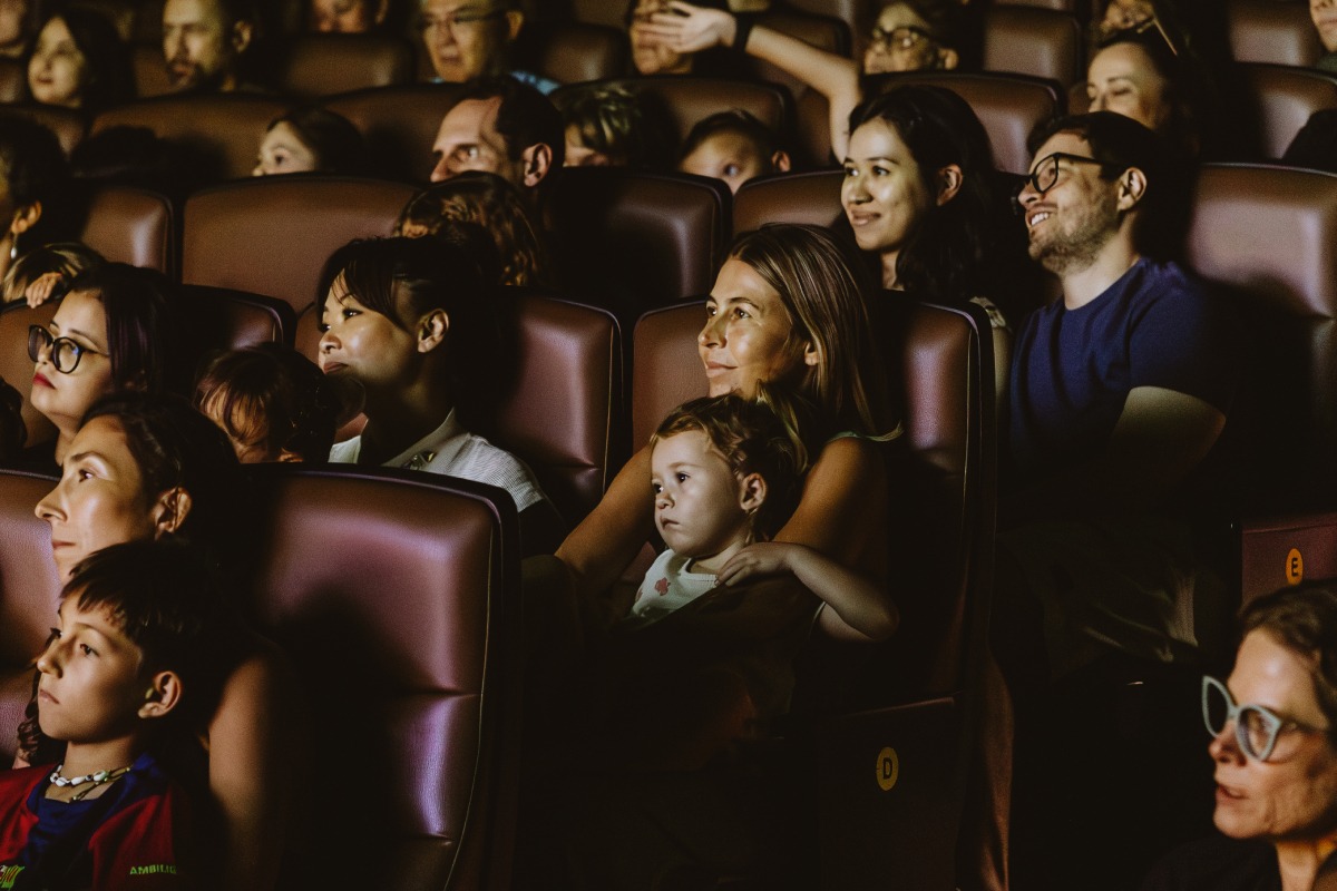 Fotografia de plateia em sala de cinema, com crianças e adultos atentos e sorridentes diante da tela.