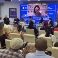 Fotografia de debate em festival de cinema na Eurásia, com participantes em roda, público atento e convidada em telão.