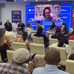 Fotografia de debate em festival de cinema na Eurásia, com participantes em roda, público atento e convidada em telão.