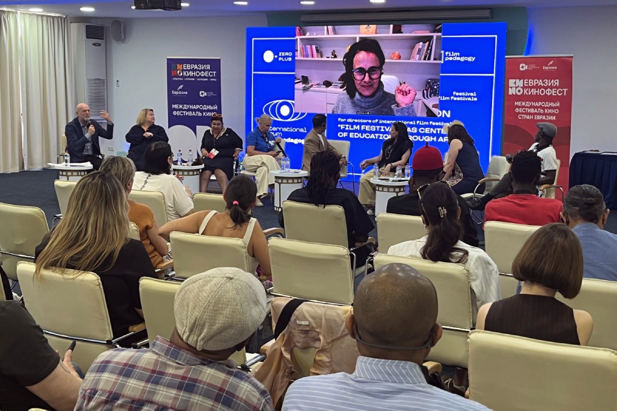 Fotografia de debate em festival de cinema na Eurásia, com participantes em roda, público atento e convidada em telão.