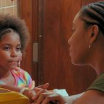 Fotografia de uma mulher negra conversando com uma menina à mesa, enquanto outra criança observa.