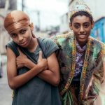 Fotografia de duas pessoas jovens em uma rua; uma cruza os braços e a outra a envolve pelos ombros.