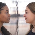 Fotografia de duas adolescentes frente a frente, uma negra de tranças e outra loira, em conversa séria.