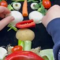 Fotografia mostra mãos organizando legumes e verduras para formar um rosto sobre uma superfície escura.