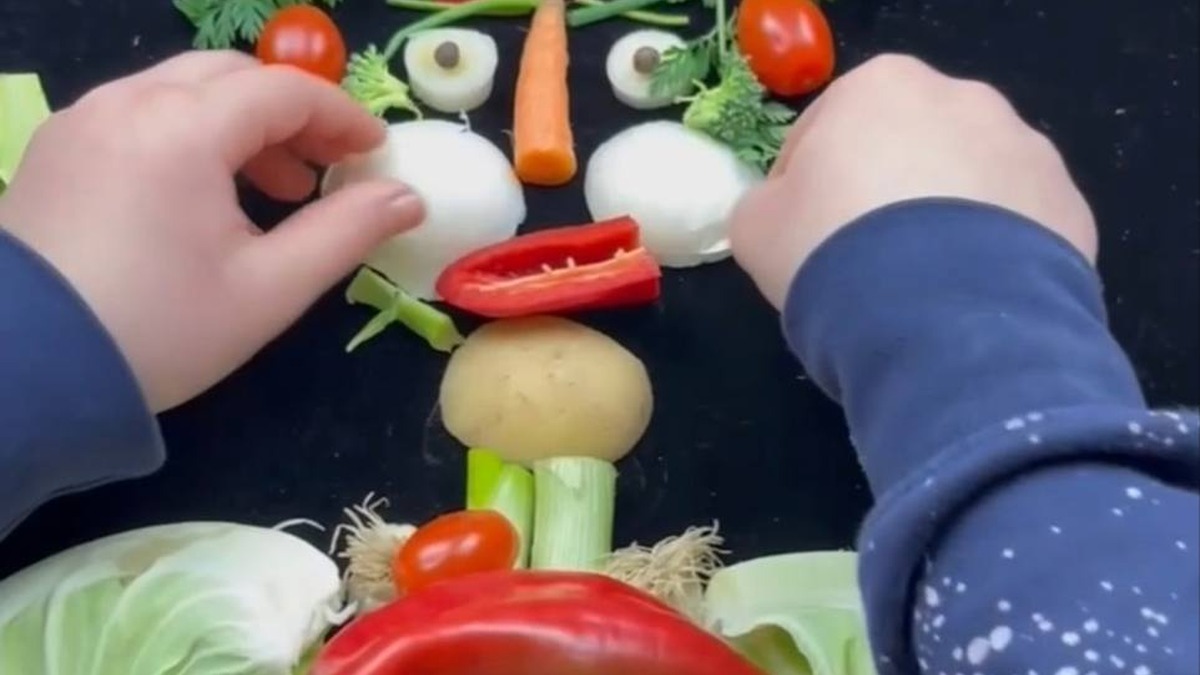 Fotografia mostra mãos organizando legumes e verduras para formar um rosto sobre uma superfície escura.