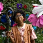 Fotografia de menino negro com óculos segurando um cajado, cercado por flores gigantes e coloridas na mata.