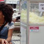 Fotografia de jovem negra sorridente sentada ao lado de um carrinho de pipoca. Texto: “pipoca, dois reais” e “não aceitamos Pix”.