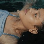 Fotografia em close de uma menina deitada, com cabelos molhados e olhar voltado para cima.