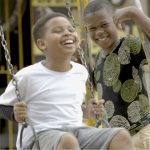 Fotografia de dois meninos negros sorrindo em um balanço; um está sentado e o outro, em pé ao lado.