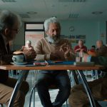 Fotografia mostra três homens idosos sentados ao redor de uma mesa, conversando em uma sala comunitária.