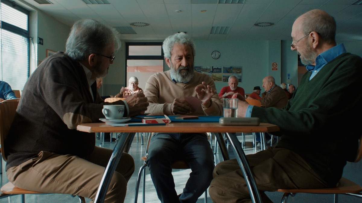 Fotografia mostra três homens idosos sentados ao redor de uma mesa, conversando em uma sala comunitária.