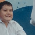 Fotografia mostra menino de camisa branca sorrindo enquanto conversa com uma mulher diante de um mural colorido.