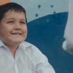 Fotografia mostra menino de camisa branca sorrindo enquanto conversa com uma mulher diante de um mural colorido.