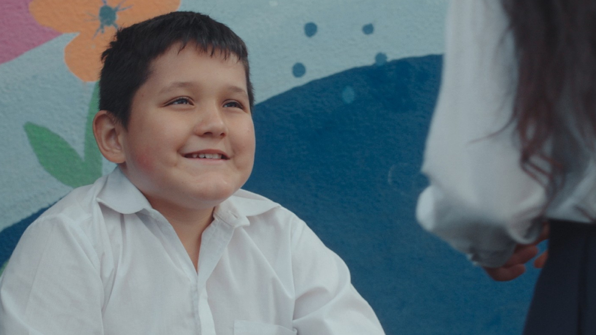Fotografia mostra menino de camisa branca sorrindo enquanto conversa com uma mulher diante de um mural colorido.