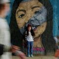 Fotografia mostra menina em pé diante de um grande mural com o rosto de uma mulher, enquanto pessoas passam ao redor.
