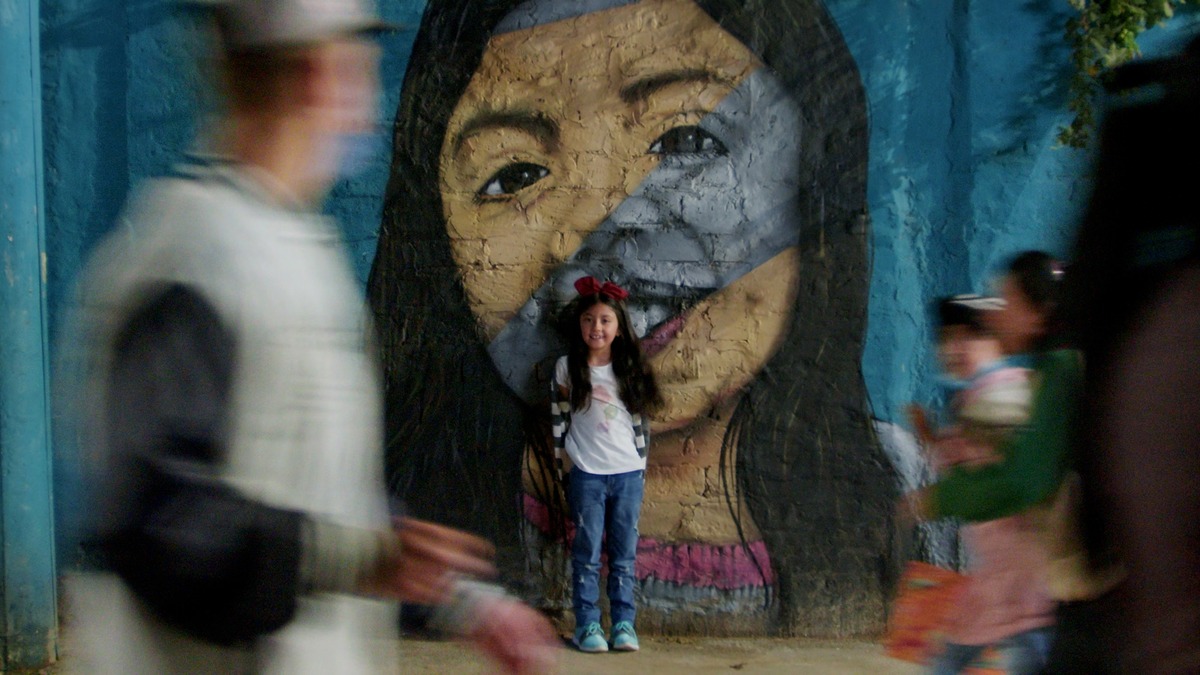 Fotografia mostra menina em pé diante de um grande mural com o rosto de uma mulher, enquanto pessoas passam ao redor.