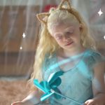 Fotografia de uma menina albina fantasiada de princesa com vestido azul, atrás de uma cortina transparente iluminada.