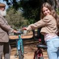 Fotografia de jovem sorridente e homem idoso de barba branca, cada um com uma bicicleta em caminho arborizado.